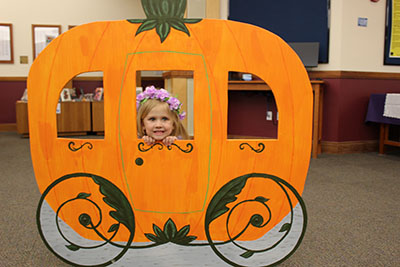 Youth with flowers in their hair peeks out of a cardboard cutout of a pumpkin carriage.