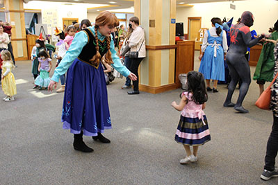 Costumed princess talks to a child in a dress in front of many costumed adults and children.